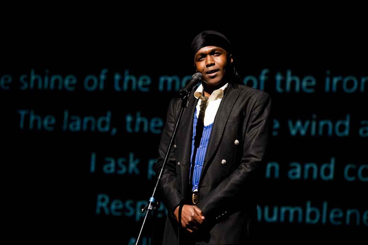 A man wearing a black jacket, white shirt, and a black headscarf speaking into a microphone on stage with a projected background of blue text.