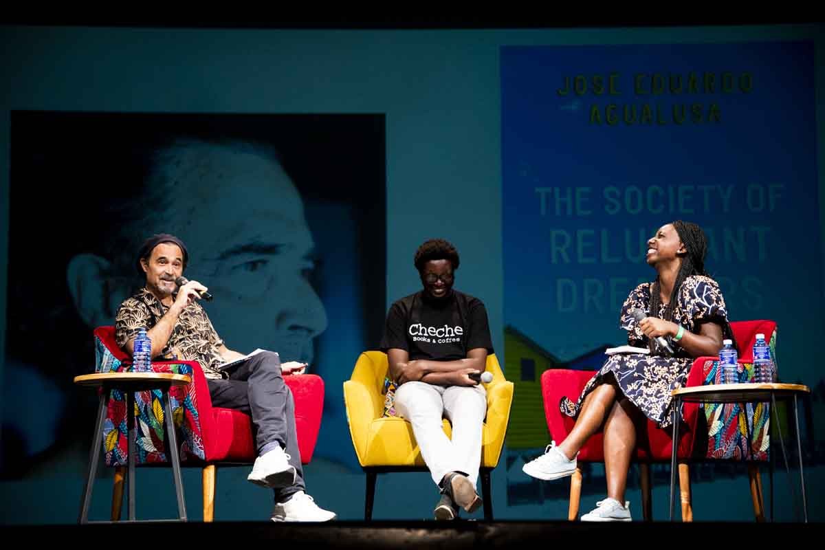 Three people sitting on colorful chairs on stage during a panel discussion, with a large book cover projected in the background. The person on the left holds a microphone and is speaking, the person in the middle is smiling and holding a microphone, 