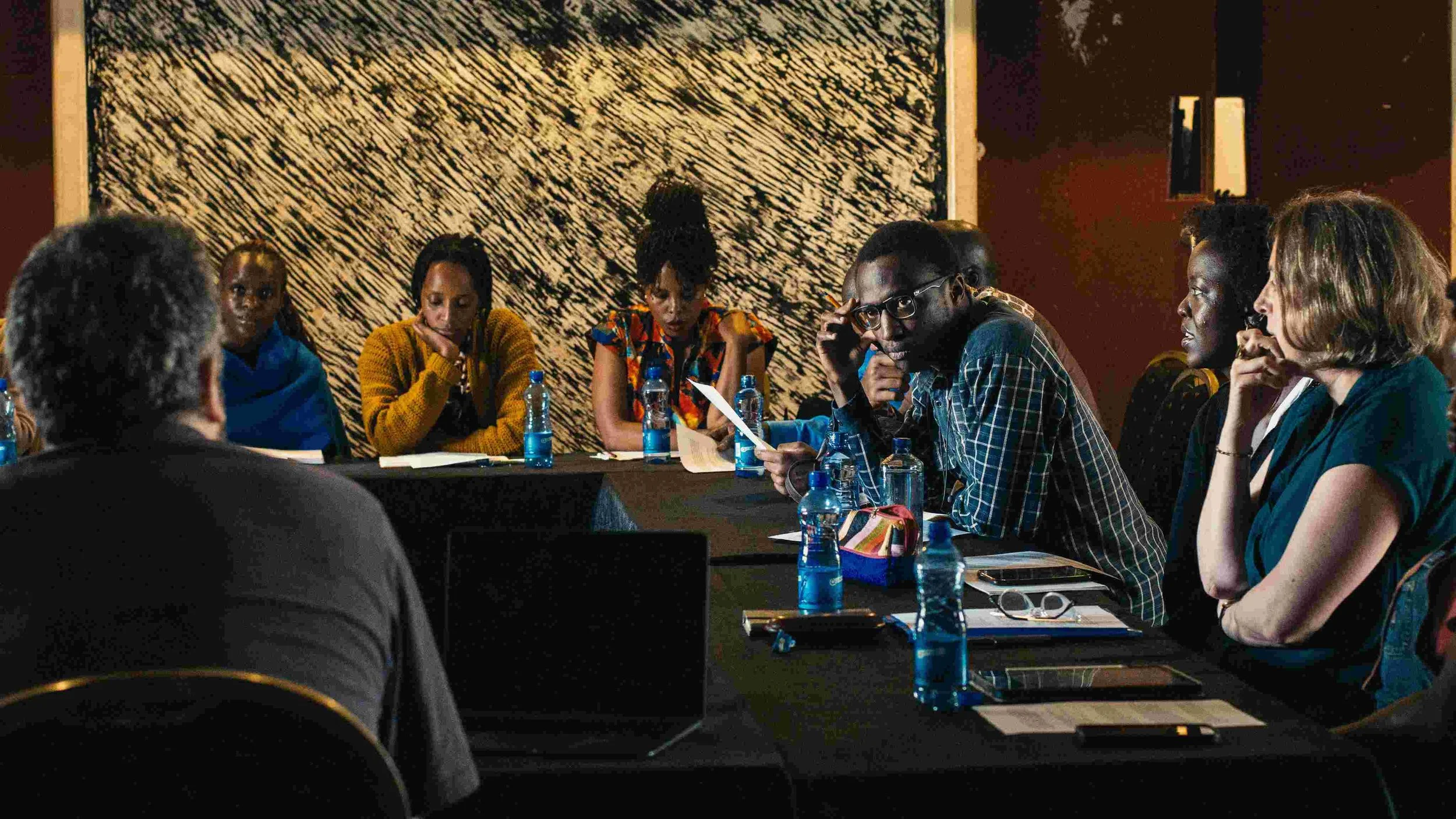 A group of people sitting around a black conference table during a meeting or discussion, with water bottles, notebooks, and electronic devices on the table, in a room with dark walls and a decorative wall hanging in the background.