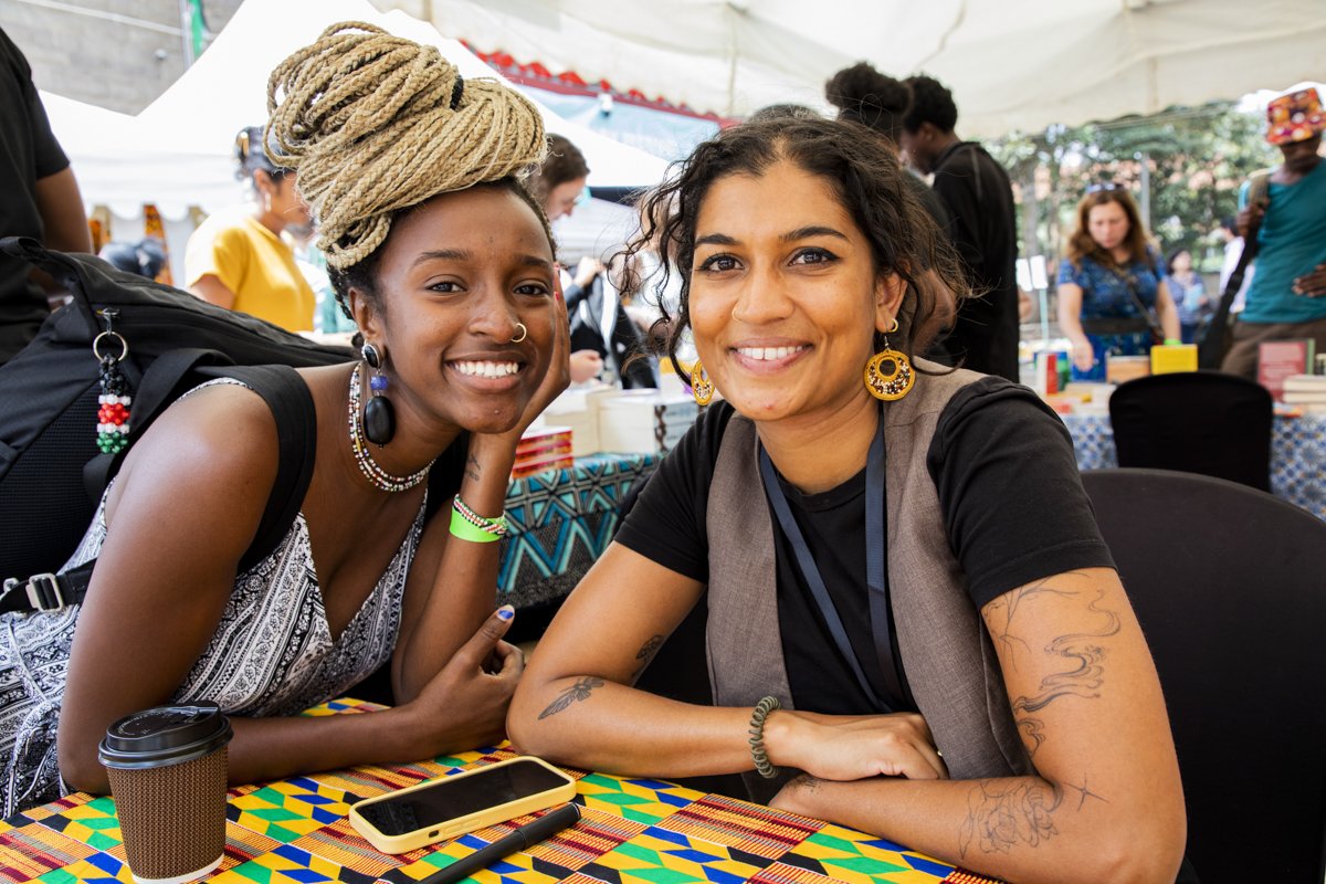 Two women sitting at a table at an outdoor event, smiling at the camera. The woman on the left has long, blonde braided hair styled in an updo, wearing a black and white patterned top and jewelry. The woman on the right has curly dark hair, wearing a