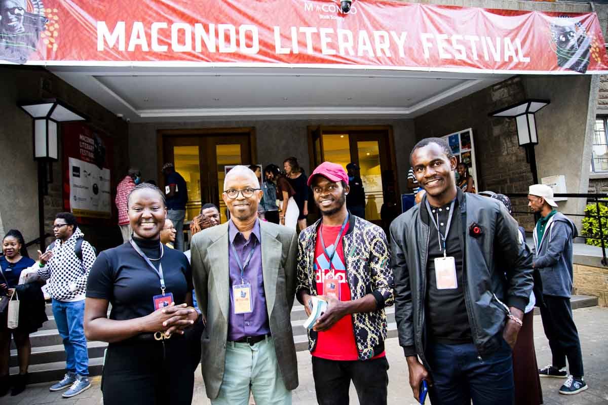 Four people smiling and standing in front of a building entrance at the Macondo Literary Festival, with others in the background, some with backpacks and wearing badges.