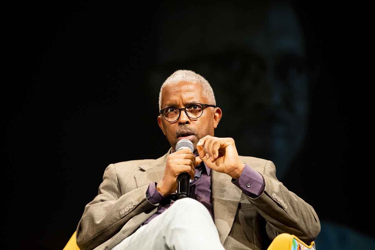 A man with gray hair, glasses, and a beard is speaking into a microphone while sitting on a yellow chair, wearing a beige blazer and purple shirt, against a dark background.