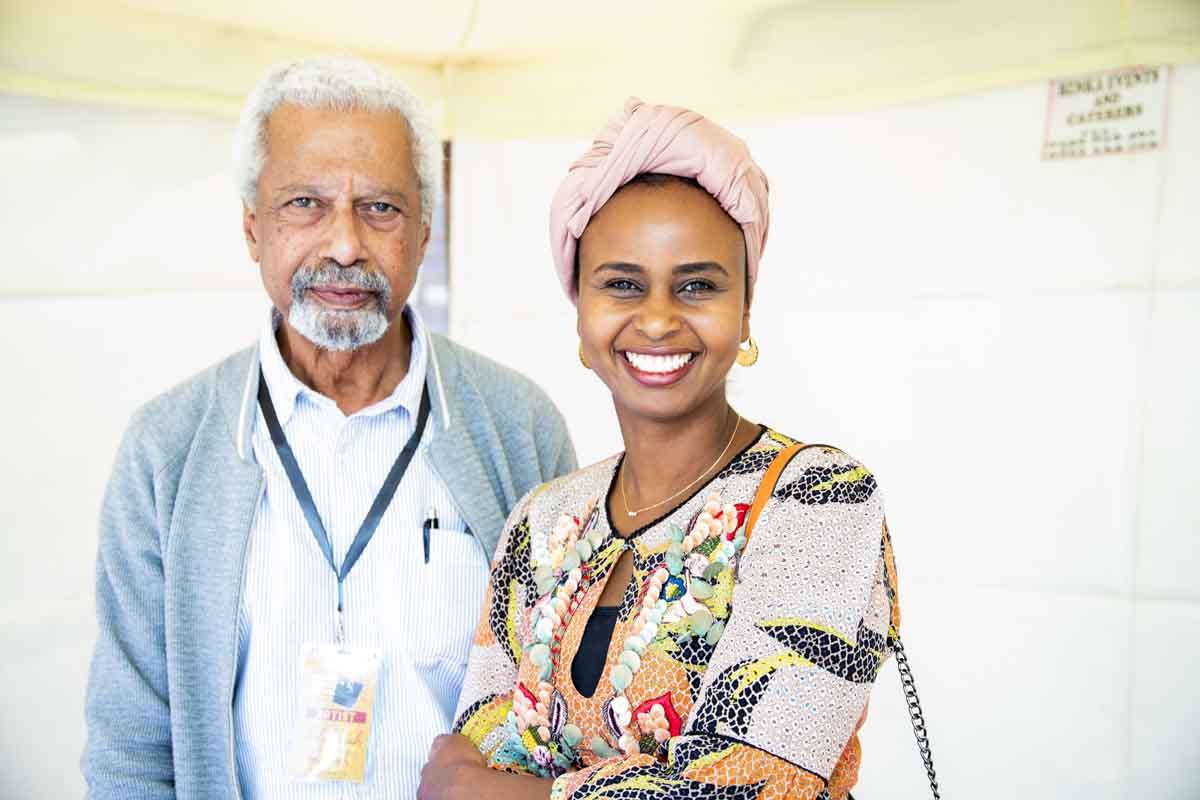 A smiling woman wearing a pink head wrap and colorful dress stands next to an elderly man with white hair, beard, and glasses, both standing in a bright indoor setting.