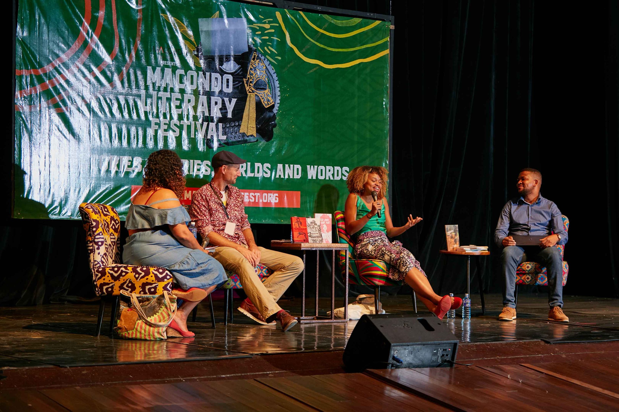 A panel of four people seated on a stage at the Macondo Literary Festival, speaking and listening to each other. The background features a large green banner with the festival's title and slogan. The stage floor is wooden, with a microphone, books, a