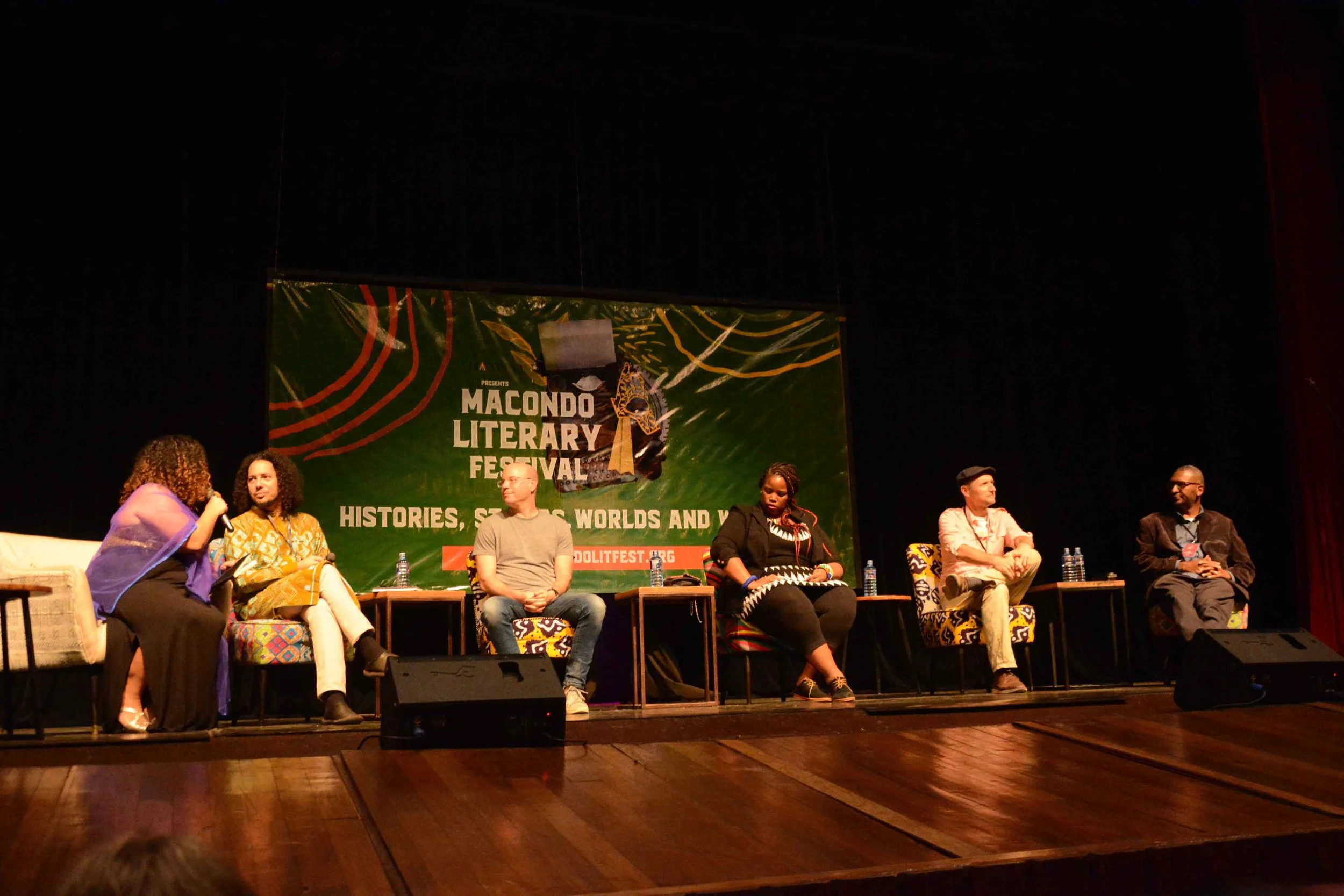 Panel discussion with six people seated on stage at Macondo Literary Festival, with a green banner in the background reading 'Macondo Literary Festival'.
