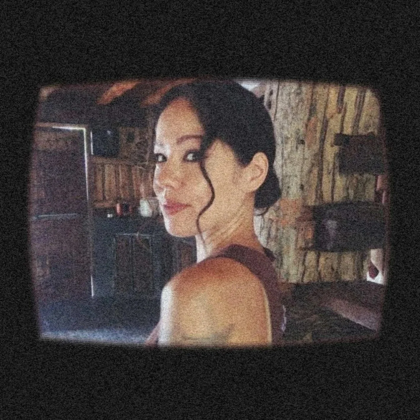 A woman with dark, wavy hair tied back, with a loose curl framing her face, looking over her shoulder at the camera. She has a calm expression, standing indoors with wooden walls and various objects in the background.