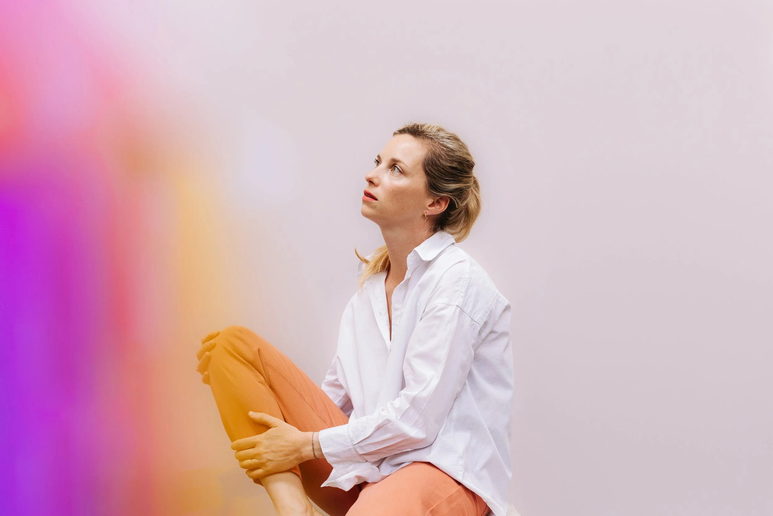 Une femme assise, tenant une jambe pliée contre sa poitrine, regardant vers le haut, avec un fond pastel dégradé.