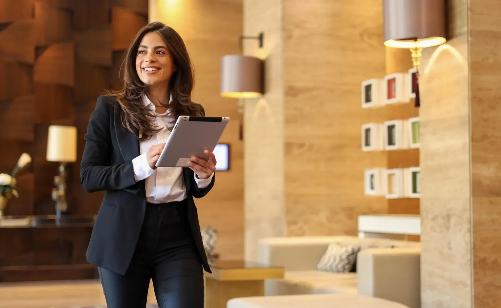 Eine lachende Frau in formeller Kleidung, die ein Tablet in den Händen hält, im modernen Hotel-LobbBereich mit Holzwänden, Buchregalen und gemütlichen Sitzecken.