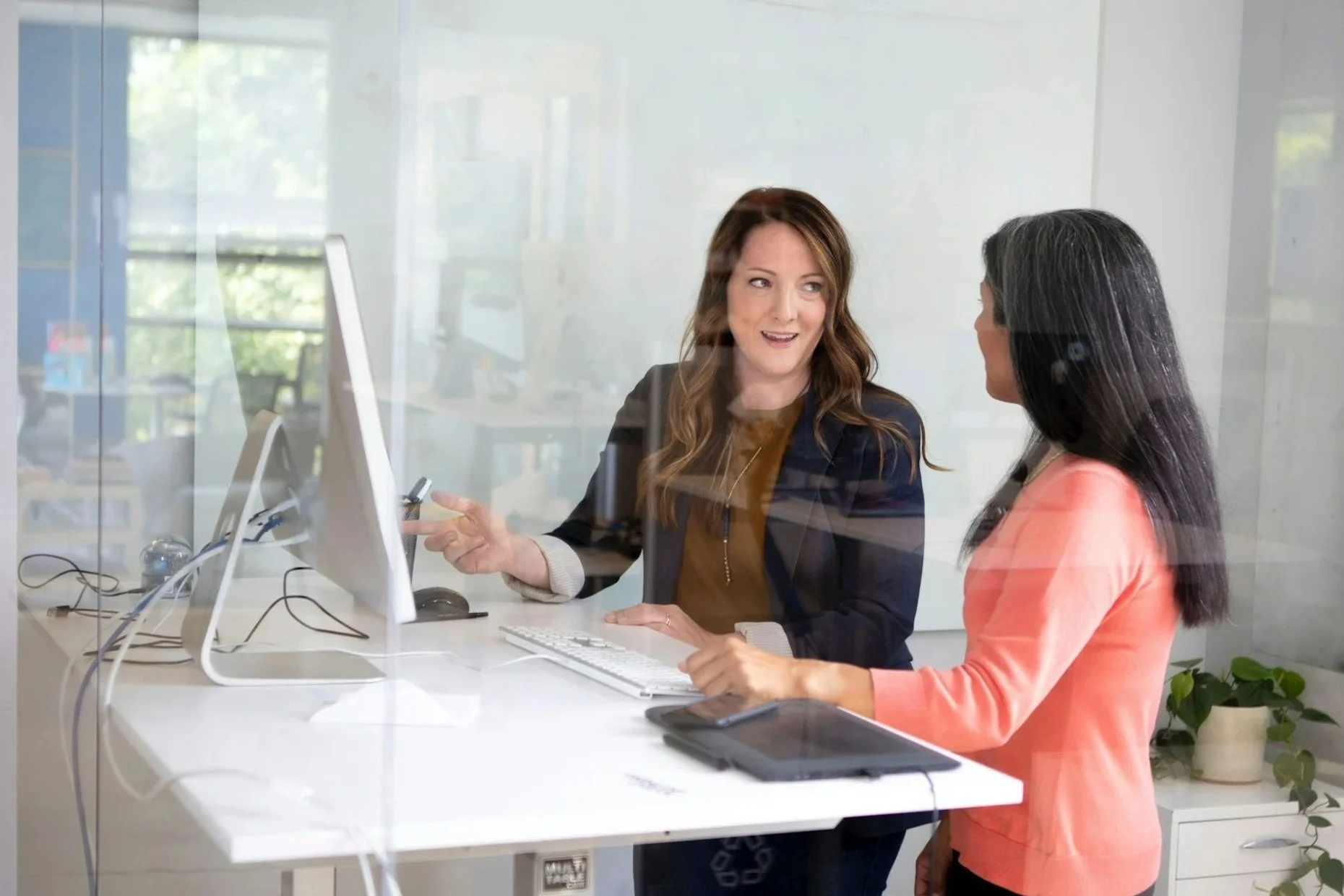 Zwei Frauen sitzen an einem Schreibtisch in einem Büro, sprechen und schauen sich an, hinter einer Glaswand.