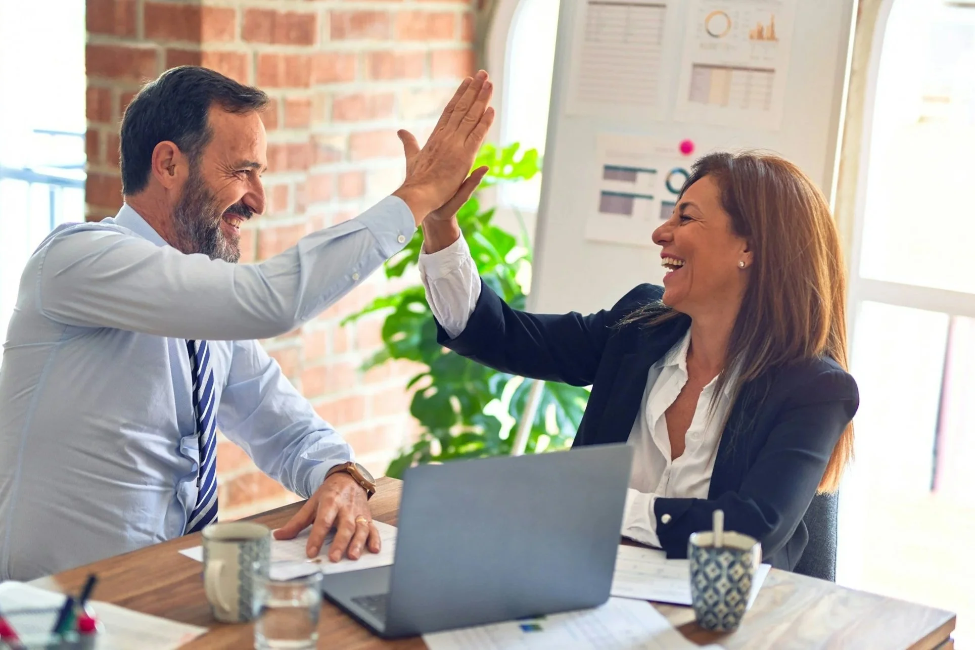 Ein Mann und eine Frau sitzen bei einem Meeting, sie feiern einen Erfolg mit einem High-Five, im Hintergrund ein Whiteboard mit Diagrammen, an ihrem Arbeitsplatz sind Kaffeetassen, Unterlagen und ein Laptop