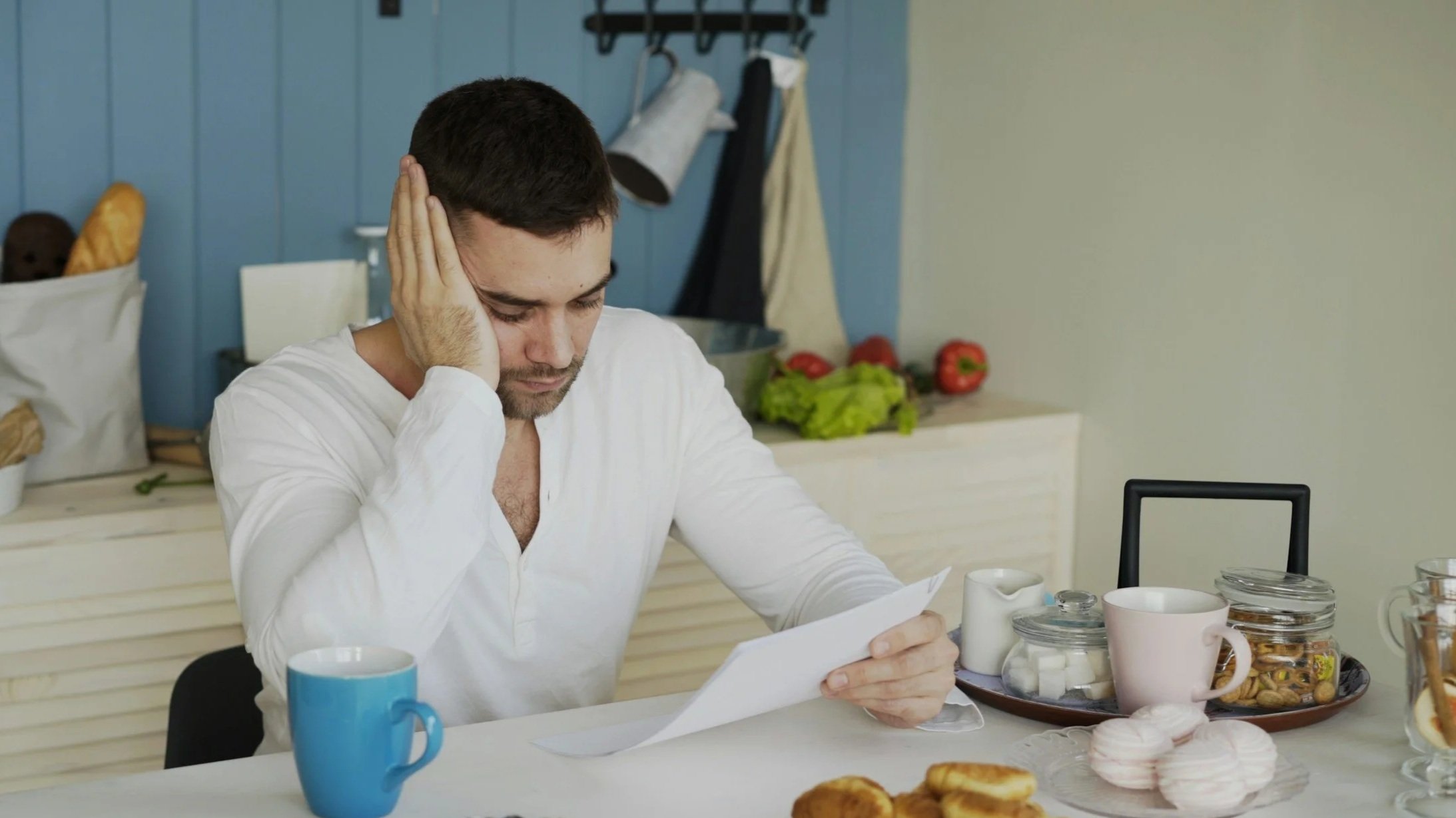 Mann liest einen Brief und sieht nachdenklich aus, während er an einem Küchentisch sitzt. Auf dem Tisch sind Kaffeetasse, Kekse, Zucker, Marmelade und andere Frühstücksartikel. Im Hintergrund sind Lebensmittel und Küchenutensilien sichtbar.