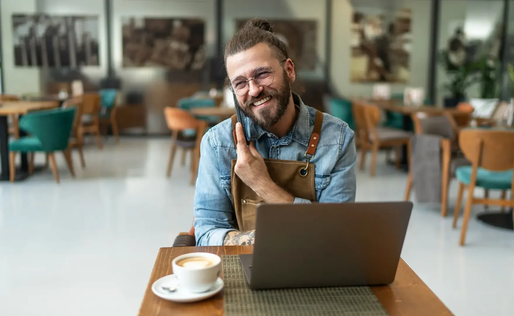 Lächelnder Mann sitzt in einem Café, spricht am Handy, hat einen Laptop vor sich und eine Tasse Kaffee auf dem Tisch.