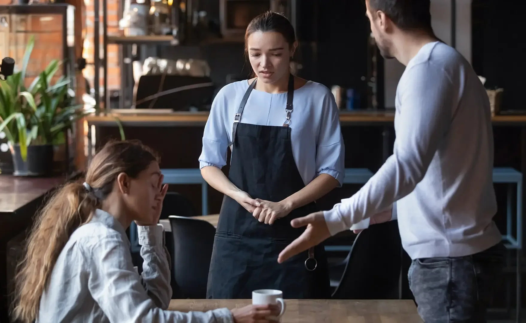 Drei Menschen in einem Restaurant, wobei eine Person traurig wirkt. Ein Mann erklärt etwas, während eine andere Frau mit einer Tasse am Tisch sitzt.