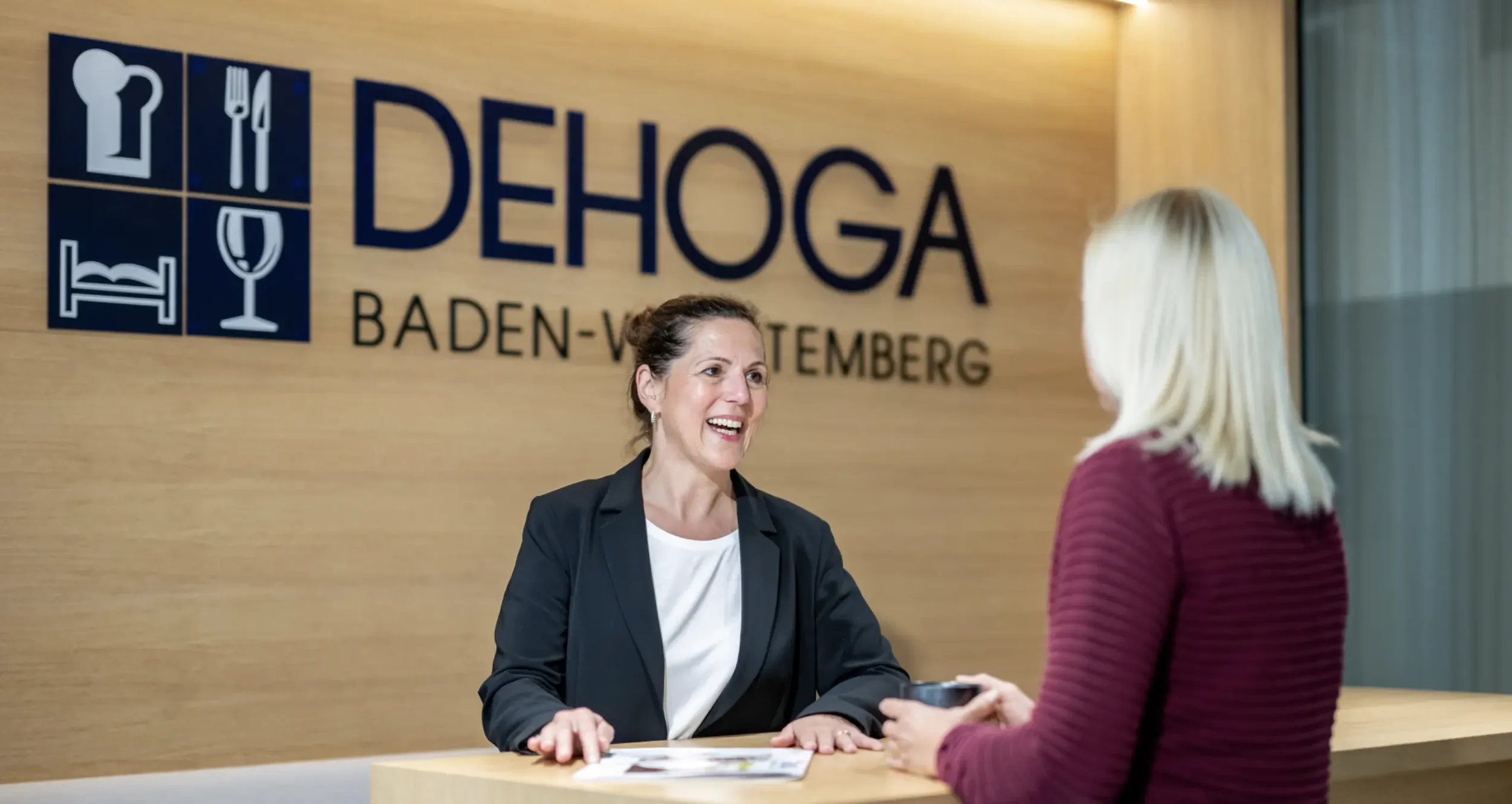 Zwei Frauen im Gespräch am Empfangstresen im DEHOGA-Büro, hinter ihnen ein Schild mit Logo und Text, das auf Baden-Württemberg hinweist.