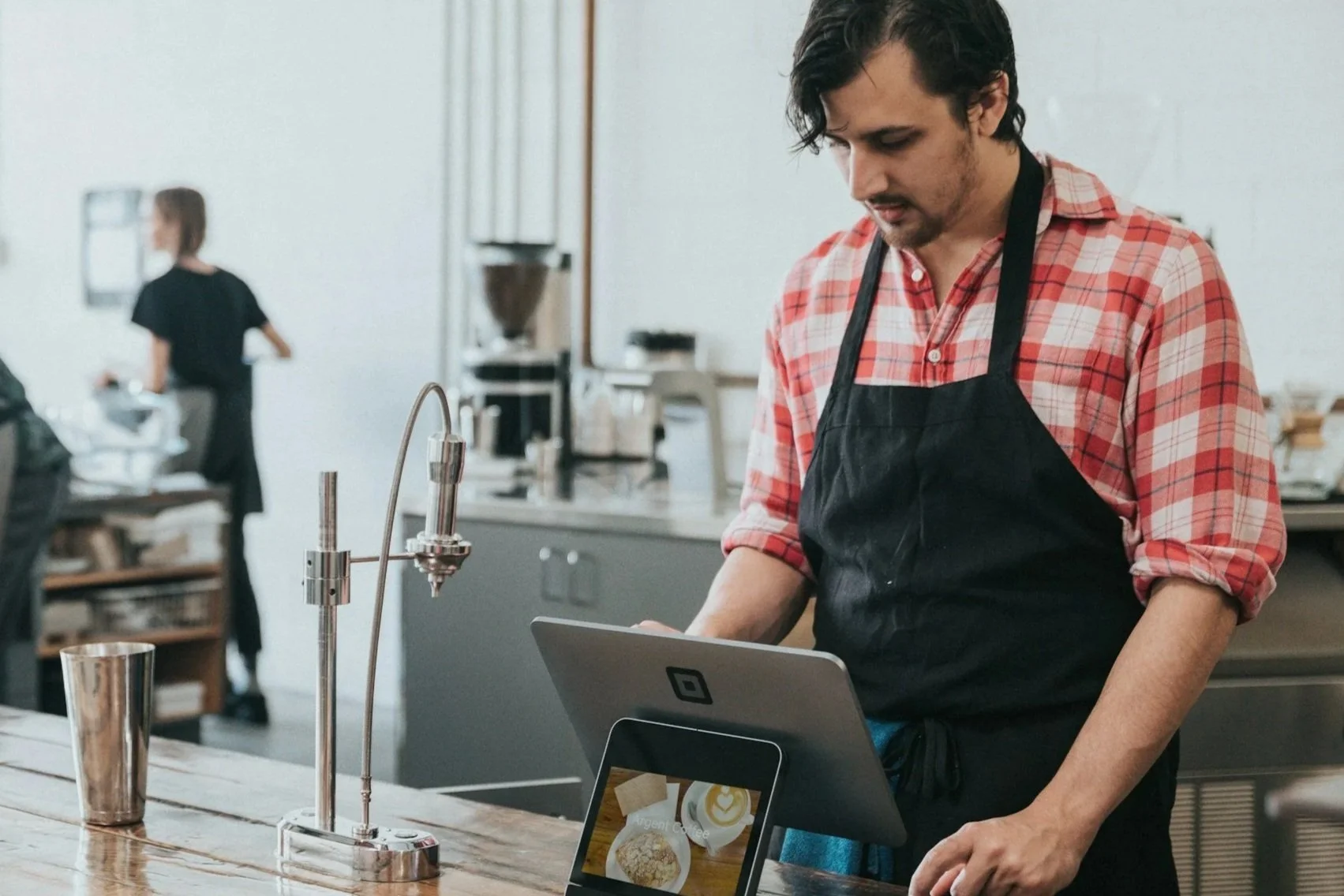Ein junger Mann arbeitet an der Kasse in einem Café oder einer Bar, trägt eine schwarze Schürze und einen rot kariertes Hemd.