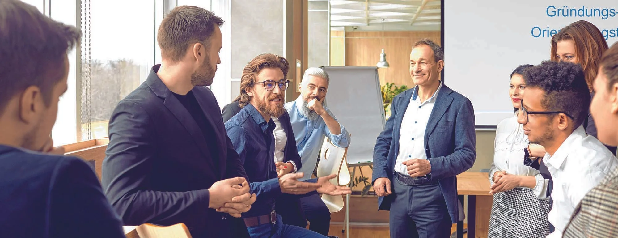 Eine Gruppe von Menschen führt eine Präsentation in einem modernen Raum mit großen Fenstern, weißem Whiteboard und Pflanzen.