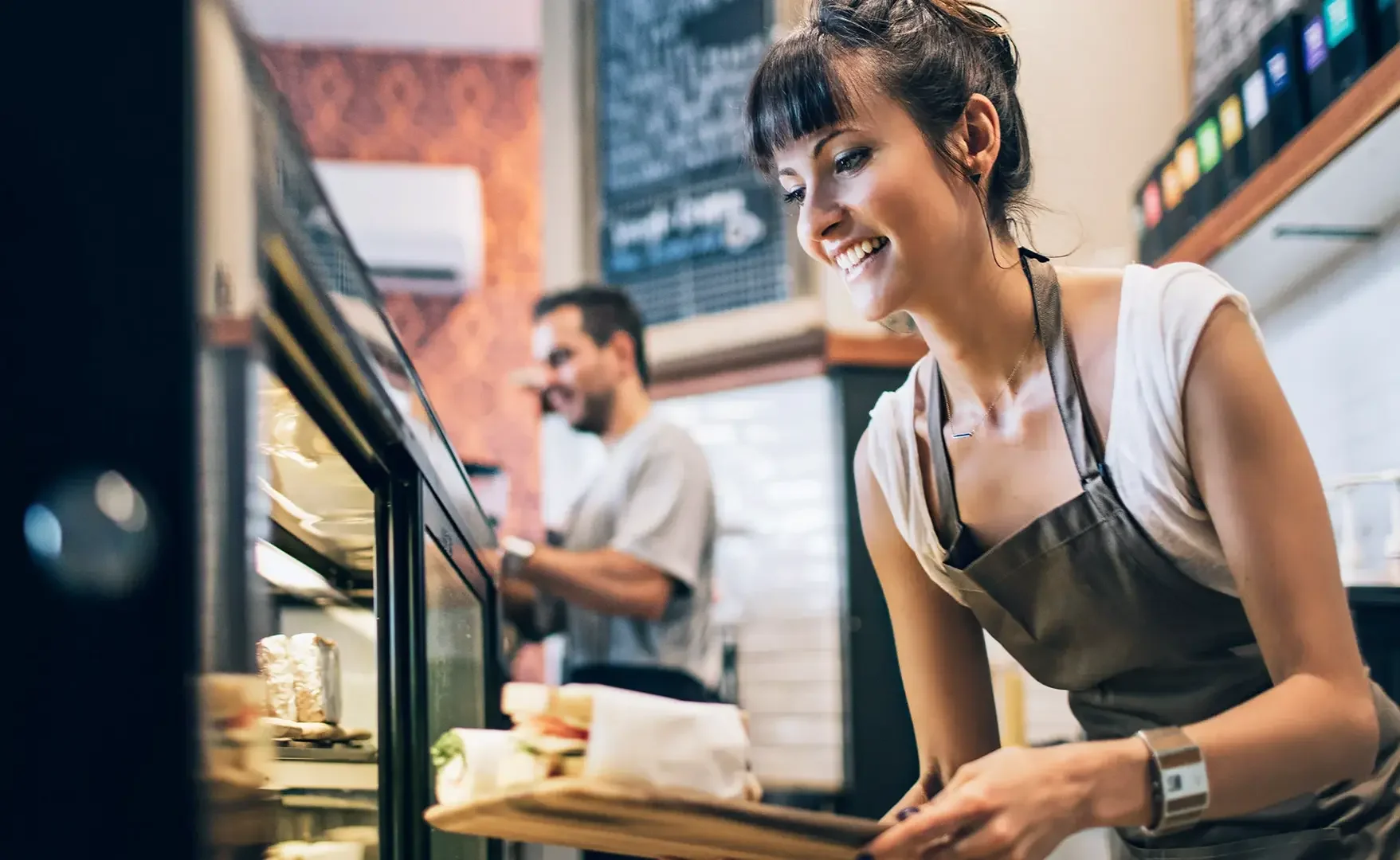 Eine junge Verkäuferin in einem Café oder Restaurant, die ein Tablett mit Sandwiches trägt, während sie hinter der Theke steht. Im Hintergrund ist ein Mann, der ebenfalls arbeitet, und ein Menübrett an der Wand.
