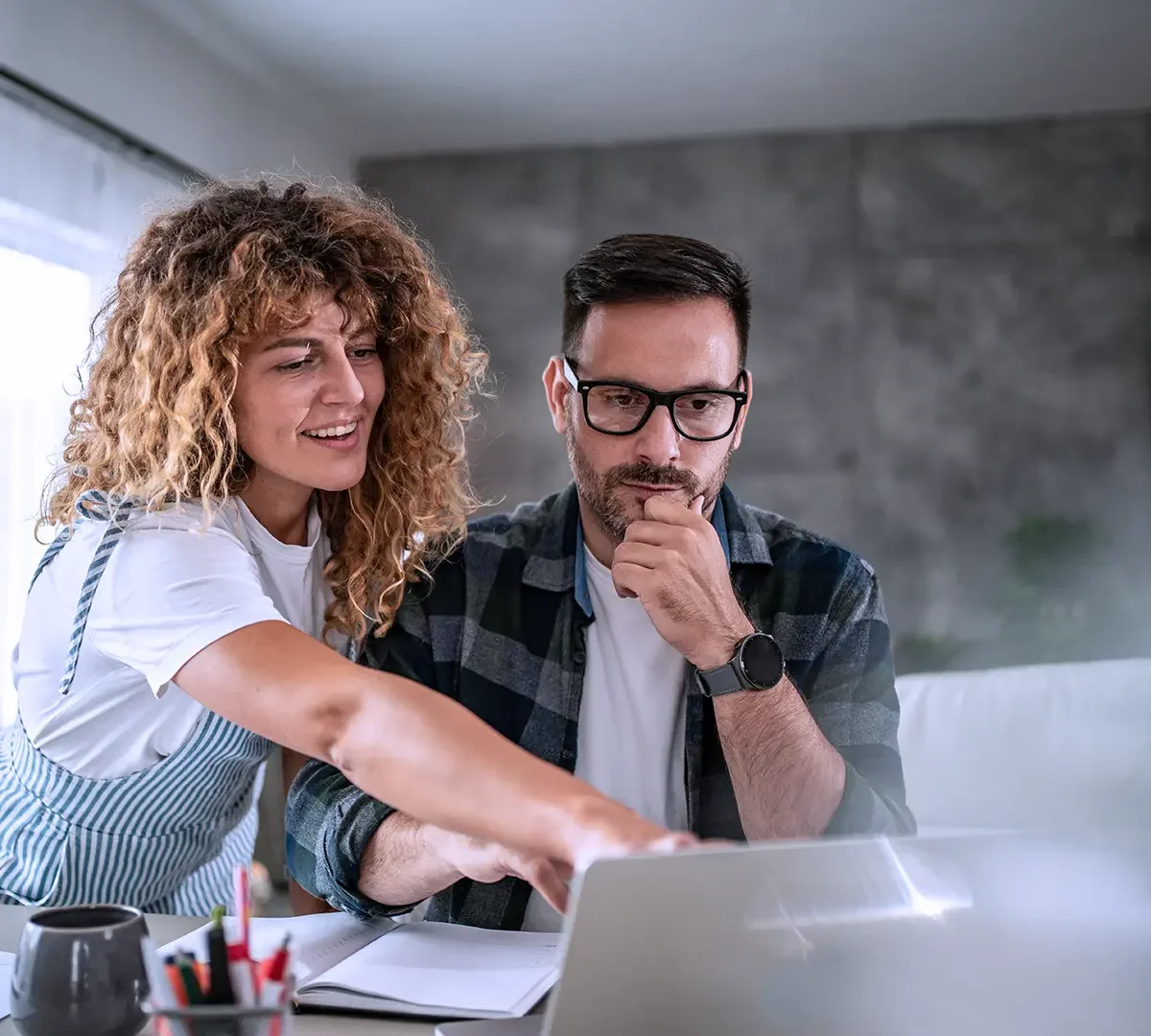 Zwei Menschen, eine Frau und ein Mann, schauen gemeinsam auf einen Laptop. Sie befinden sich in einem modernen Raum, die Frau zeigt auf den Bildschirm, der Mann denkt nach.