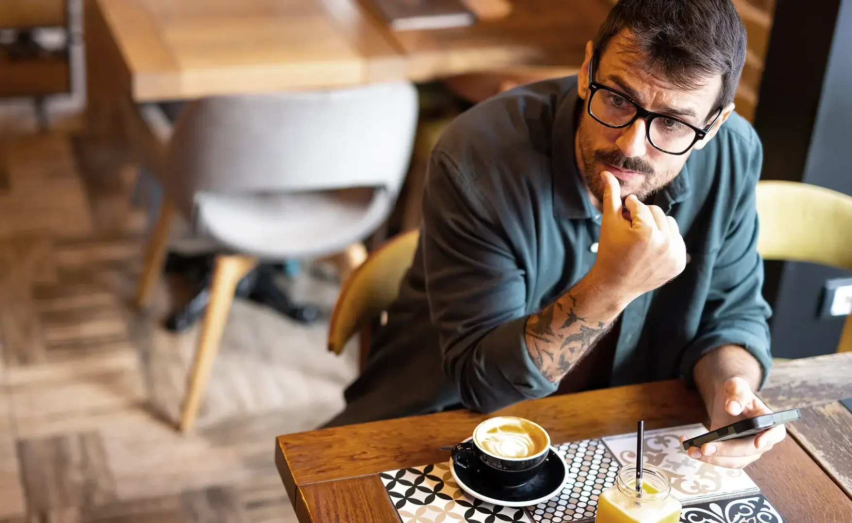 Ein Mann mit Brille, Tattoos am Arm, sitzt an einem Holztisch in einem Café, schaut auf sein Smartphone, mit Kaffee und einem Glas Orangensaft vor ihm.