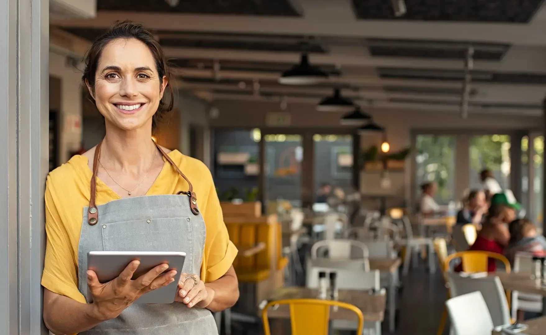 Eine lächelnde Frau in gelber Bluse mit grauer Schürze hält ein Tablet in einem Restaurant.