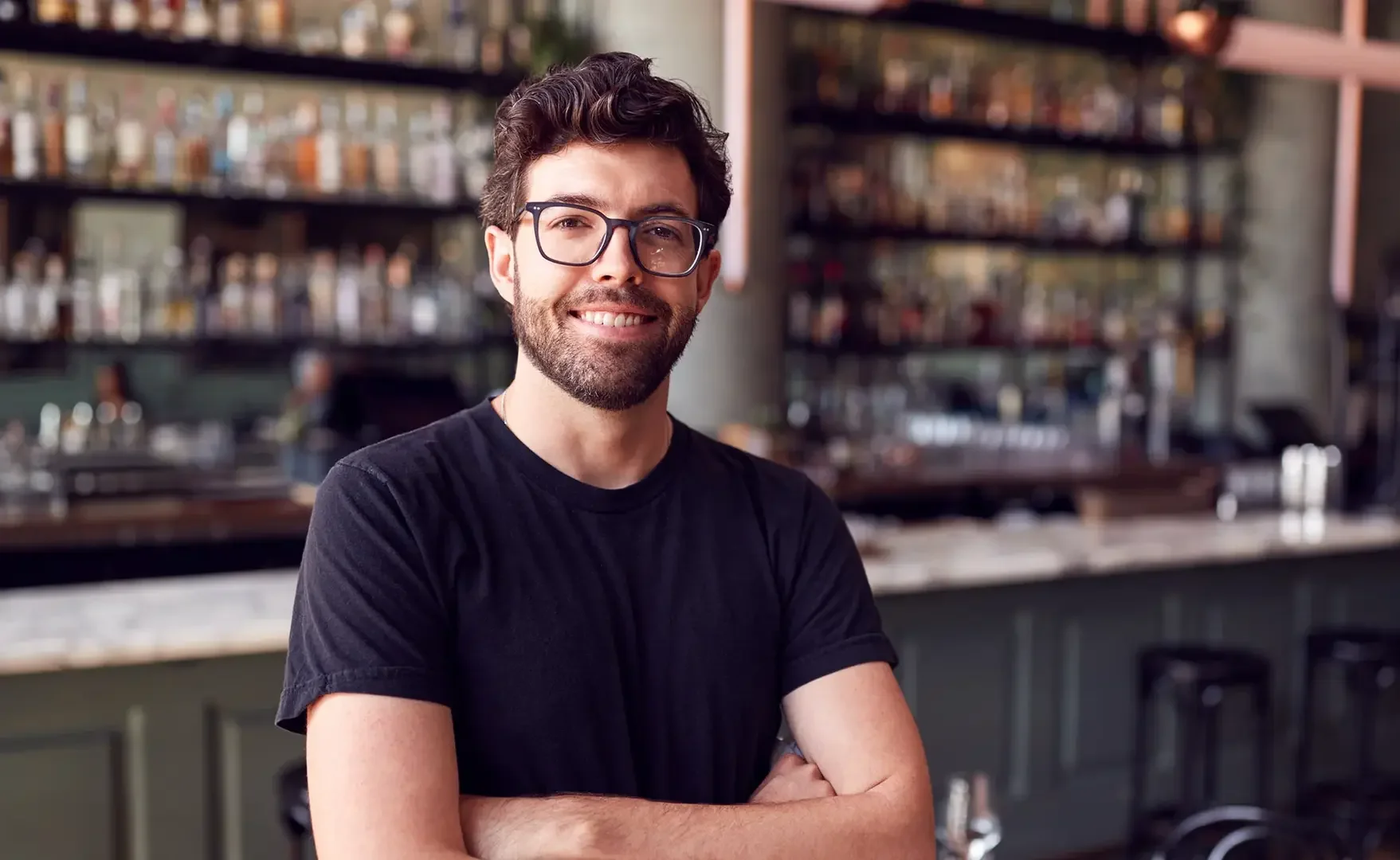 Ein junger Mann mit dunklem Haar, Bart, Brille und schwarzen T-Shirt lächelt an einem Barhintergrund in einem Restaurant oder Café.