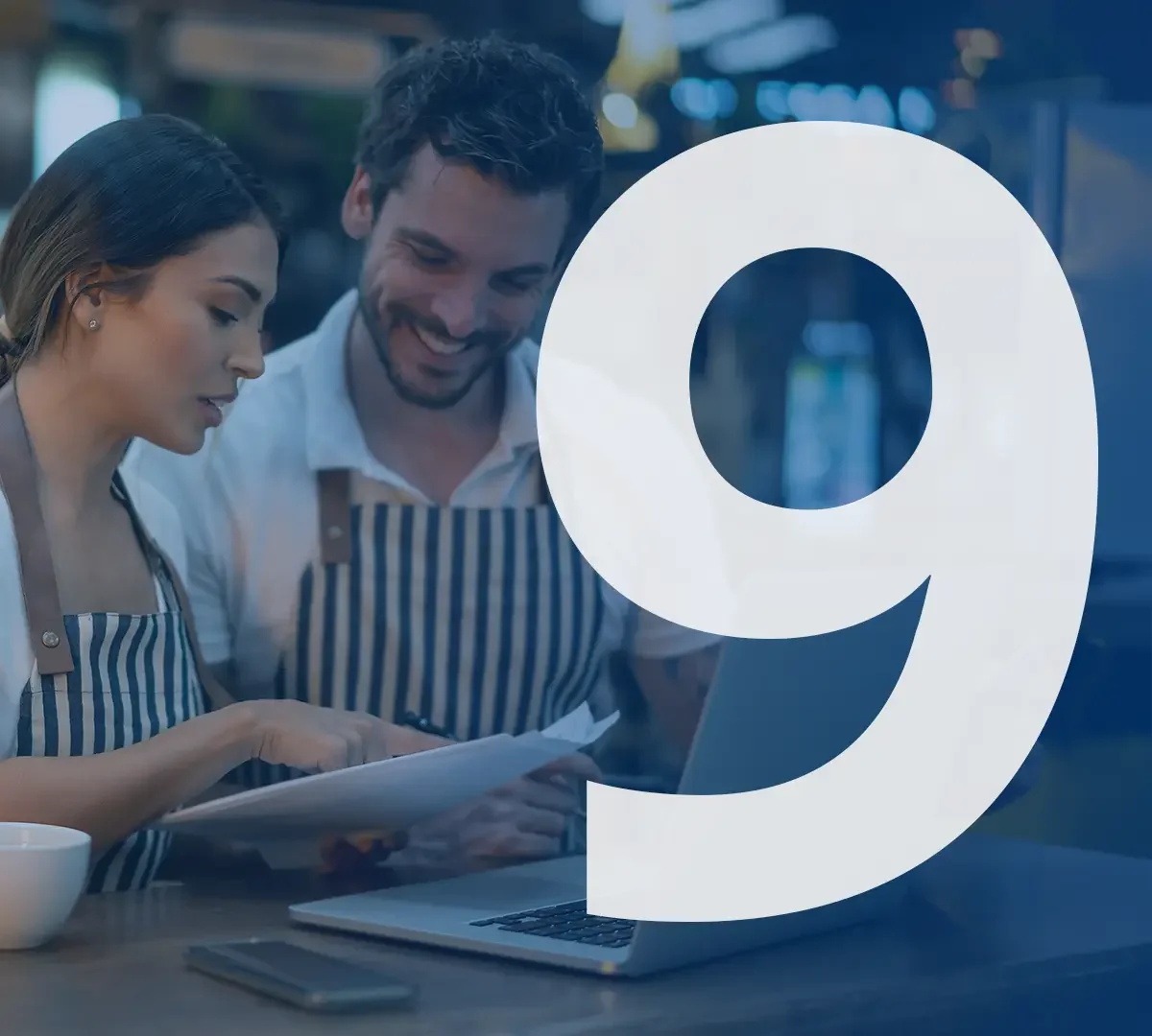 Zwei Menschen, ein Mann und eine Frau, in einem Café mit Laptops und Papieren, lachen und schauen gemeinsam auf ein Dokument, während sie eine angenehme Unterhaltung führen. Im Vordergrund eine große weiße Zahl 9.