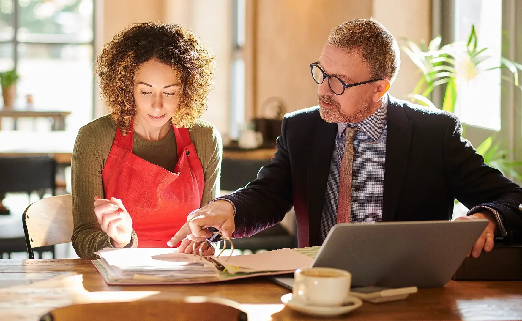 Zwei Personen, eine Frau mit Locken und braunen Haaren und ein Mann mit kurzen Haaren und Brille, sitzen an einem Tisch in einem Café, schauen in ein Fotobuch und unterhalten sich.