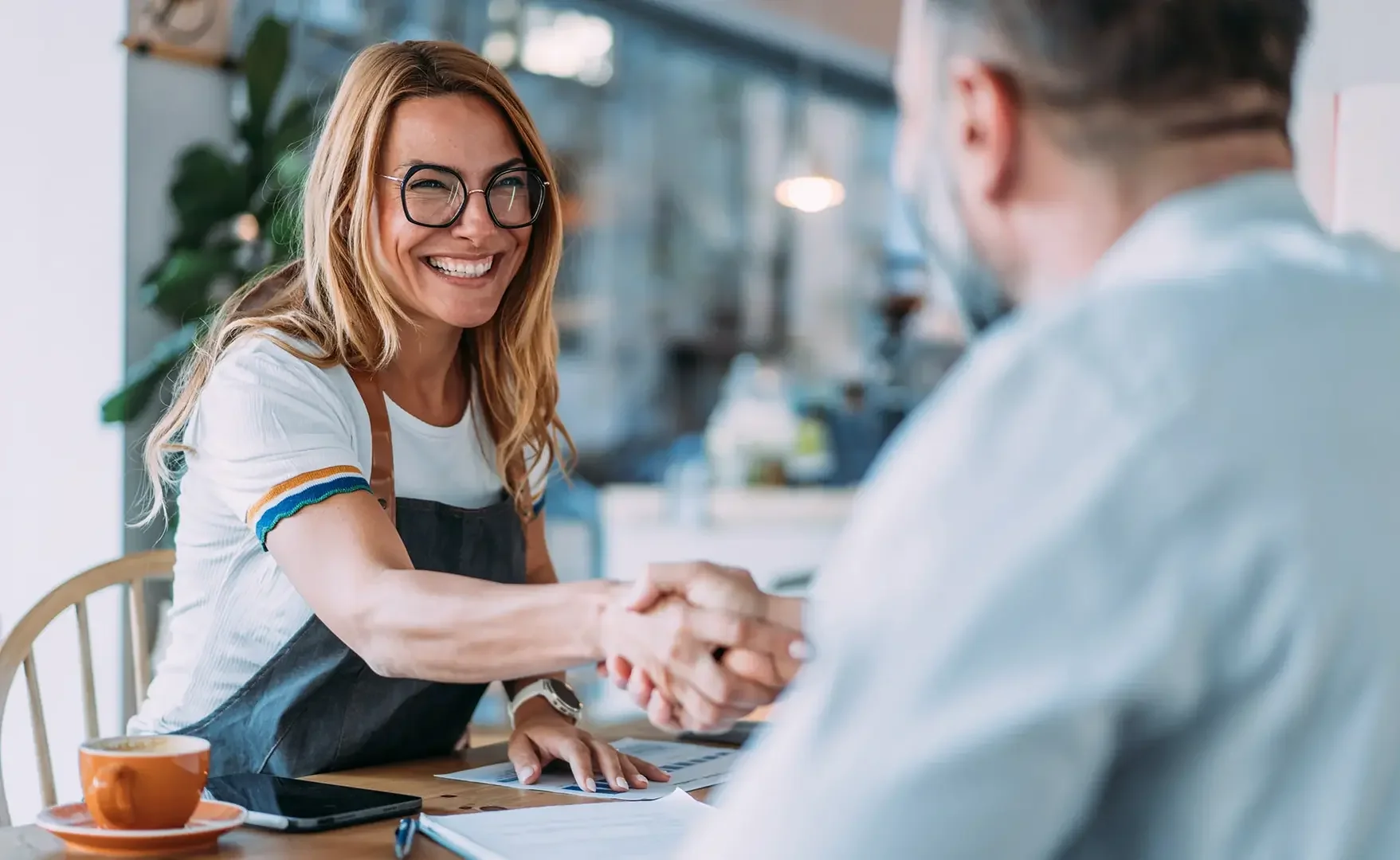 Eine lächelnde Frau beim Händeschütteln mit einem Mann in einem Café oder Restaurant. Im Hintergrund sind Pflanzen und das Innere des Lokals sichtbar.
