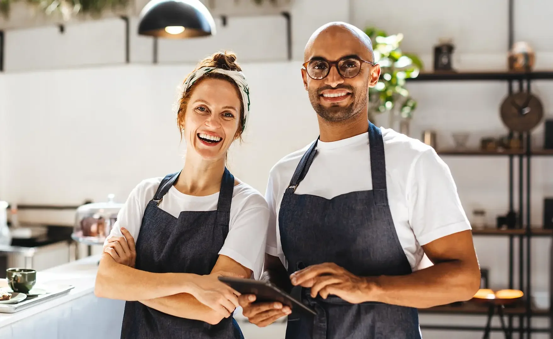 Zwei freundliche Baristas, eine Frau und ein Mann, stehen in einer modernen Kaffee-Bar. Sie tragen schwarze Schürzen und lächeln in die Kamera, während sie ein Tablet halten.