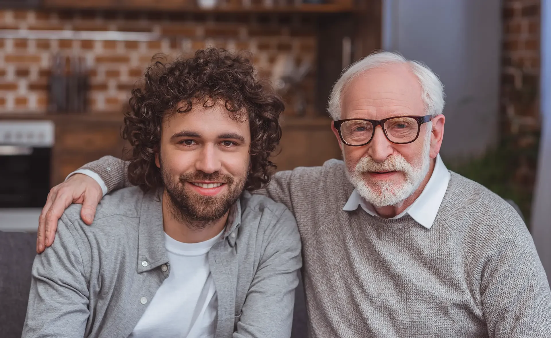 Ein älterer Mann mit weißen Haaren und Brille sitzt neben einem jüngeren Mann mit dunklem, lockigem Haar. Beide lächeln und schauen in die Kamera, im Hintergrund eine Küche mit Backsteinwand.