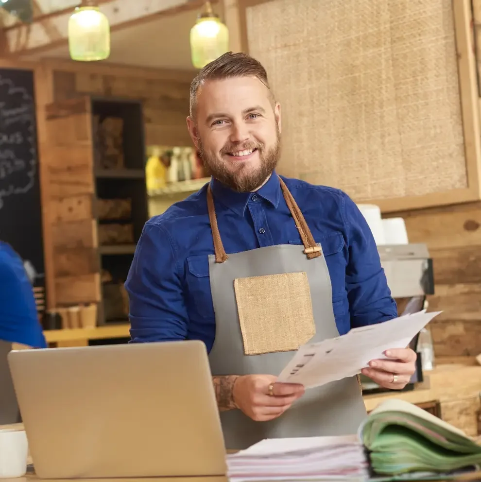 Ein lachender Mann mit Bart, blauer Hemd und einem grauen Schürze hält Papiere in der Hand. Im Vordergrund befindet sich ein Laptop auf einem Tisch. Die Umgebung ist eine gemütliche, holzverkleidete Küche oder Café. Der Mann wirkt freundlich und professionell.