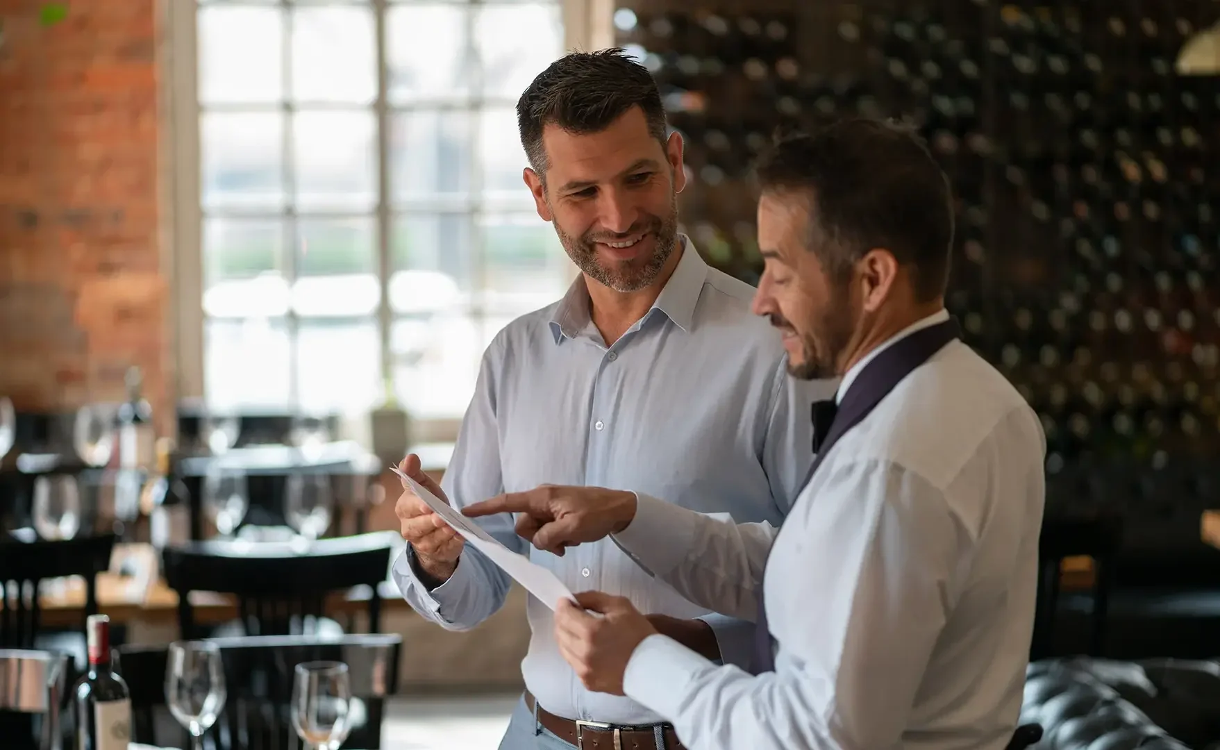 Zwei Männer im Gespräch, einer zeigt auf eine Speisekarte in einem Restaurant mit viel natürlichem Licht.