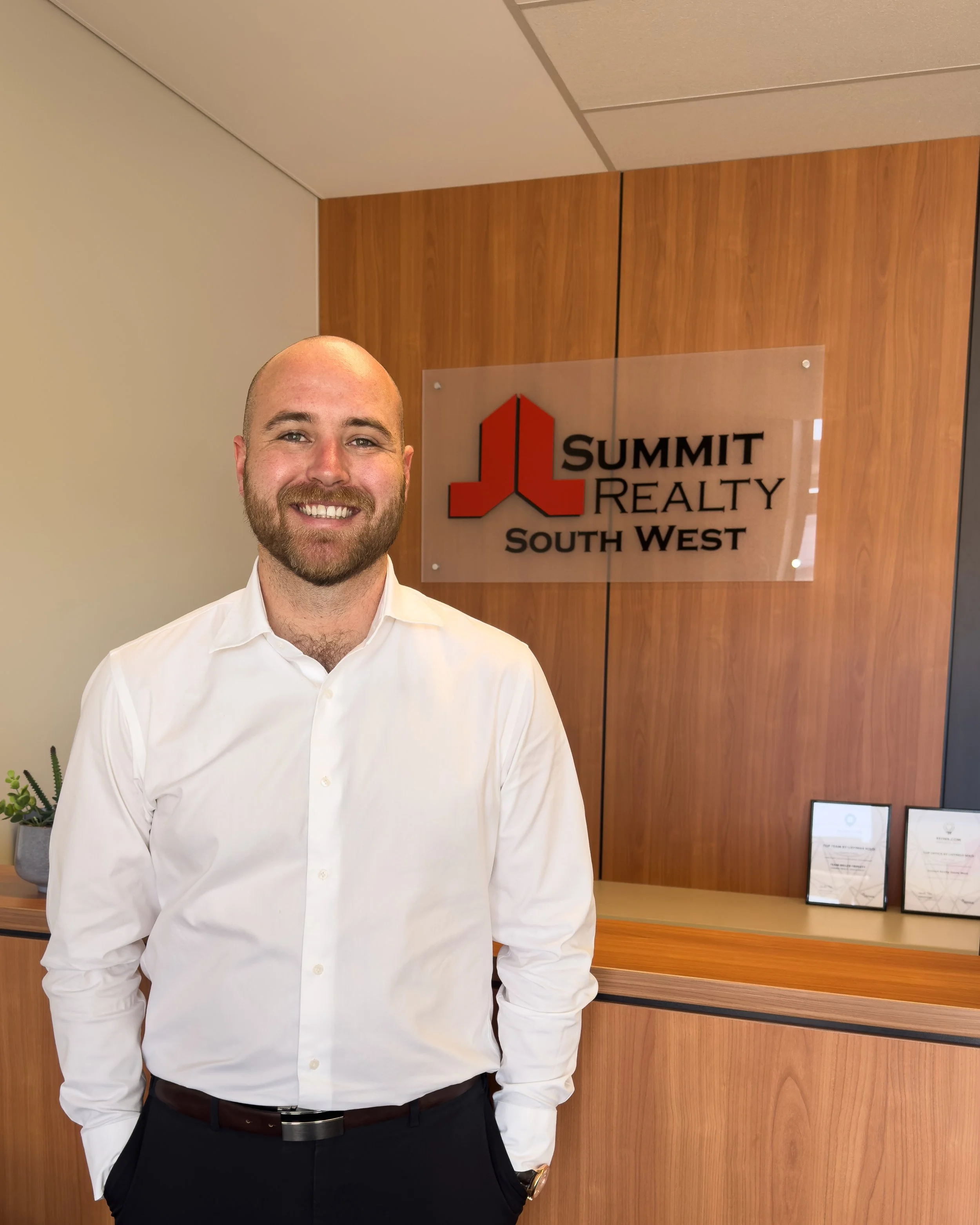 Ben Wood, A smiling man with a beard and a white shirt stands in front of a Summit Realty South West sign at a reception desk, with awards or certificates in frames on the counter and a small potted plant nearby.