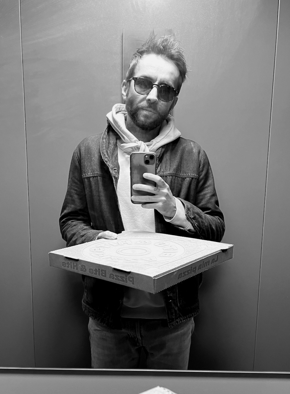Autoportrait en noir et blanc d’un homme barbu portant des lunettes de soleil, un sweat à capuche et une veste en jean, tenant une boîte à pizza et un smartphone devant un miroir.