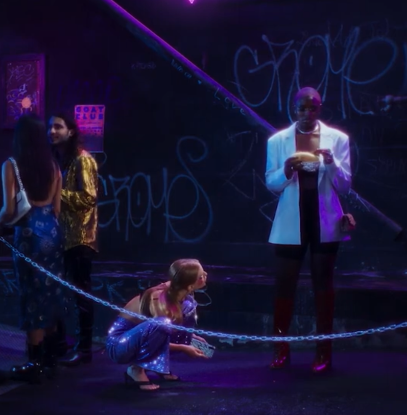 Scène de spectacle plongée dans une lumière violette, avec plusieurs personnes costumées devant un mur couvert de graffitis et derrière une chaîne.