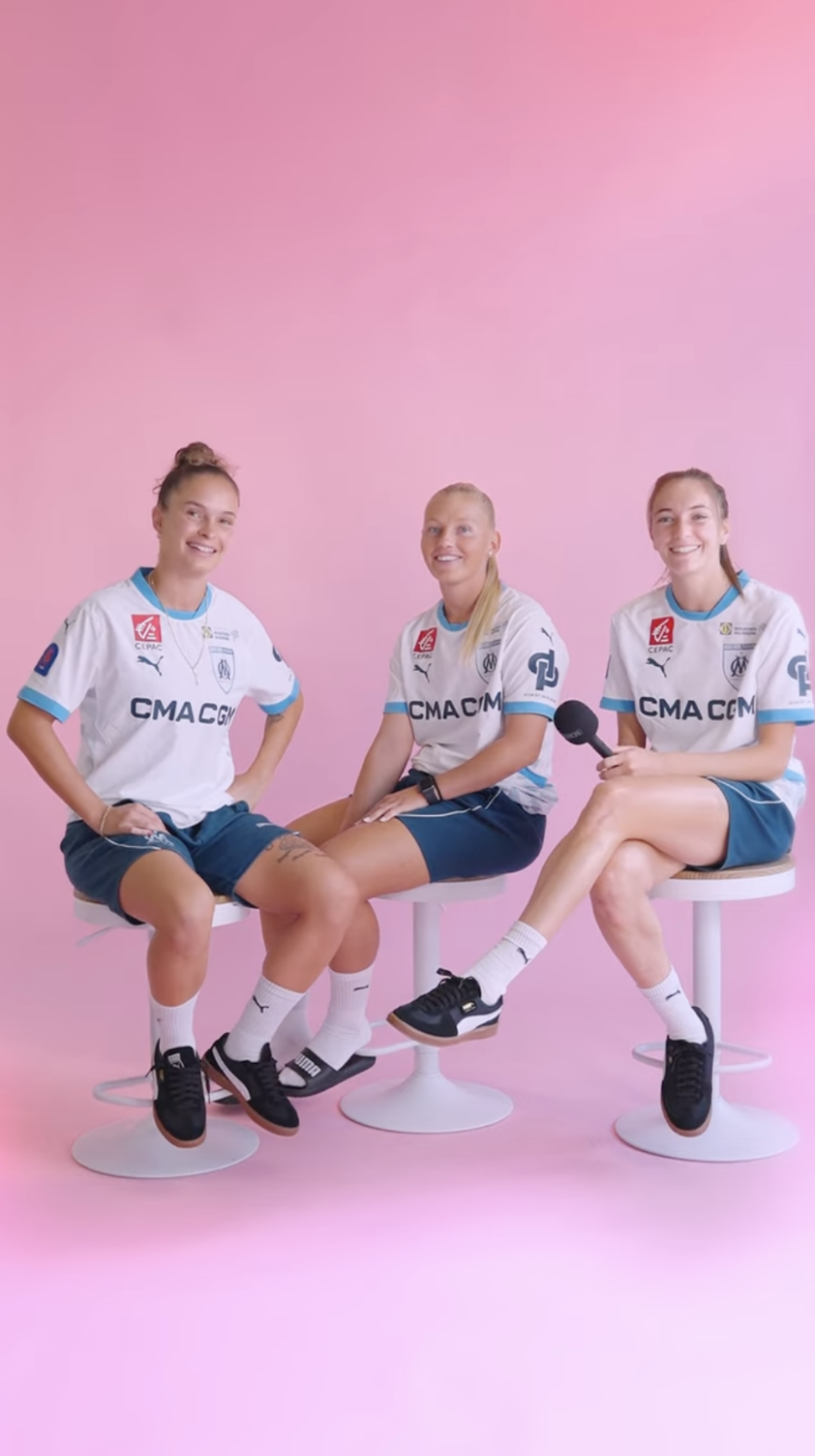 Trois femmes en tenue de sport assises sur des tabourets blancs, souriantes, contre un fond rose, l'une d'elles tient un micro.