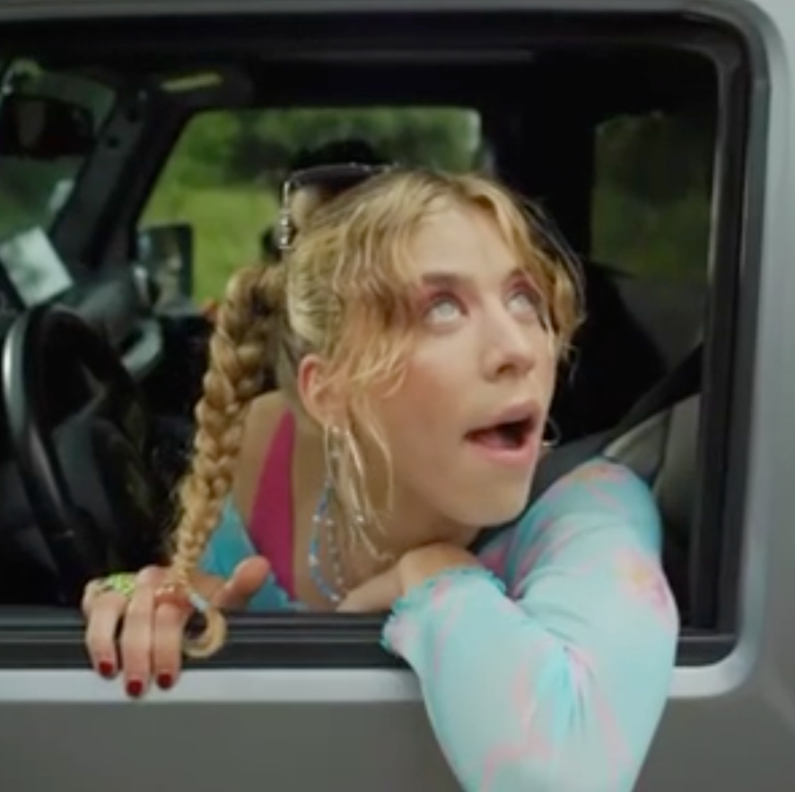 Femme blonde tressée se penchant par la fenêtre ouverte d’un véhicule, l’air surprise.