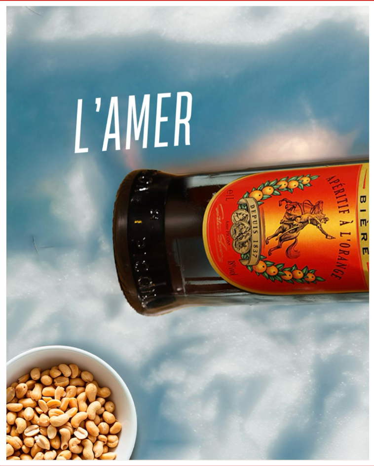 Bouteille de bière d’abbaye belge posée sur la neige, avec un bol de cacahuètes et le texte « L’amer ».