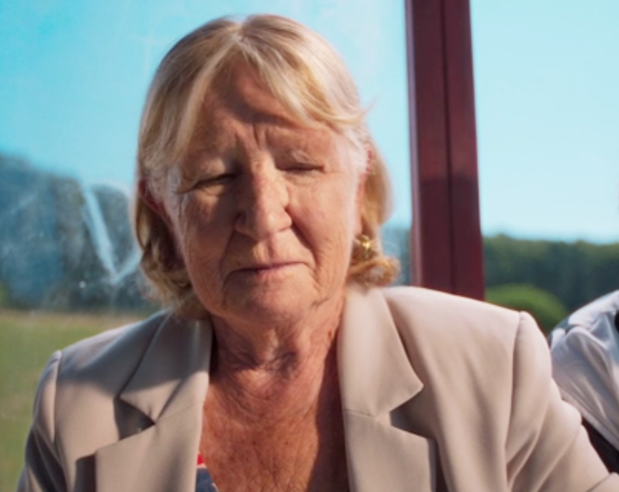 Portrait rapproché d’une femme âgée aux cheveux blonds, portant un blazer clair, devant un paysage extérieur.