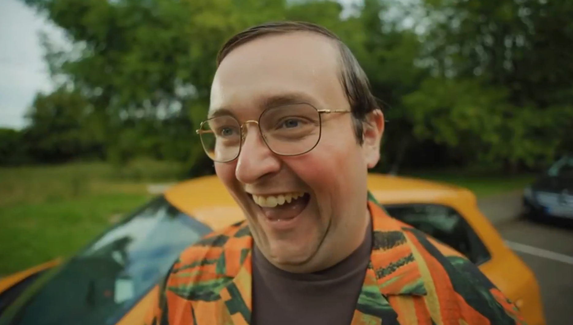 Homme souriant portant des lunettes et une veste colorée devant une voiture orange, dans un environnement verdoyant.