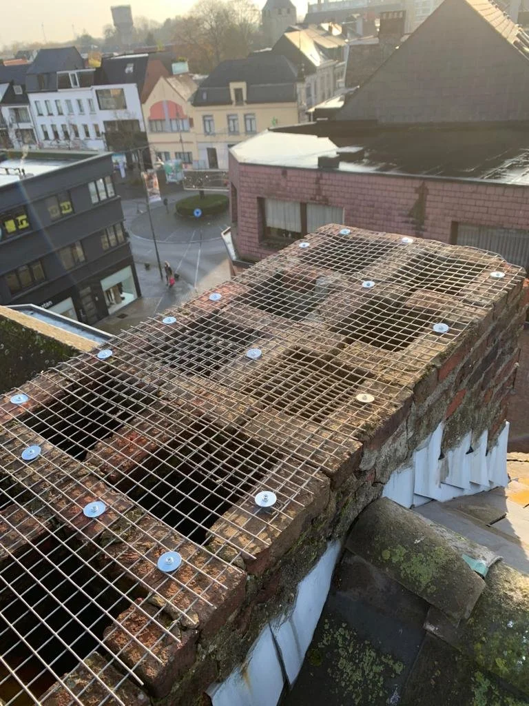 Een oude bakstenen schoorsteen met een metalen rooster en uitsparingen, op een daken van een gebouw, met een stad en huizen op de achtergrond.