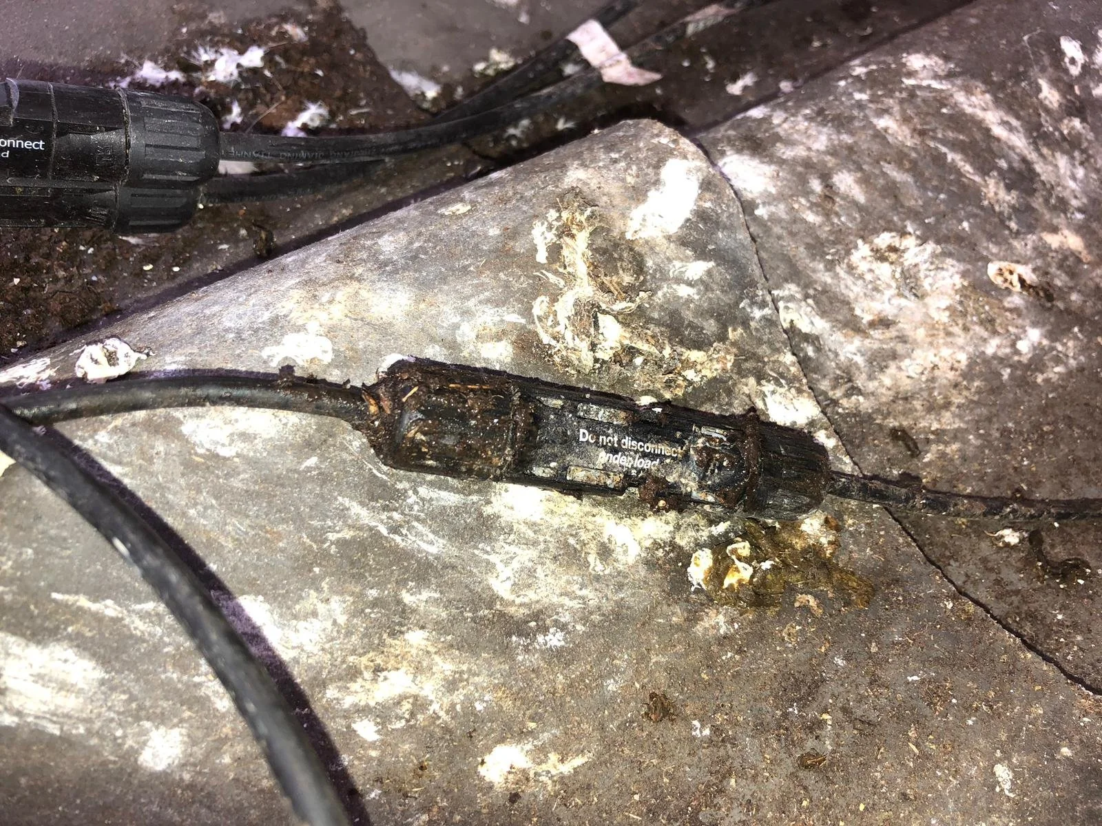 Een elektrische kabel met een beschadigde en roestige connector ligt op een stenen vloer.