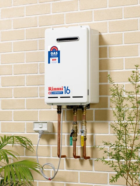 A Rinnai hot water heater mounted on a beige brick wall, with gas and water connections, nearby green plants and an electrical outlet.