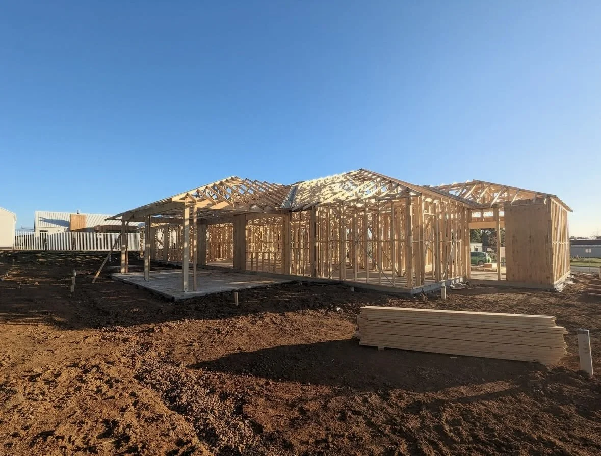 The wooden framework of a house under construction on a dirt lot, with a clear blue sky in the background.