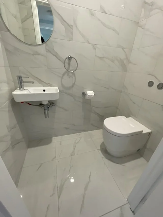 Small modern bathroom with marble walls and floor, a white wall-mounted sink, a round mirror, a wall-mounted toilet, and a towel ring.