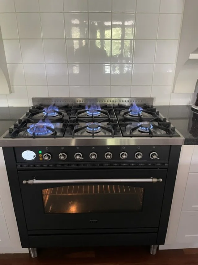 A black gas stove with six burners, all lit with blue flames, and an oven below with a glass door, in a kitchen with white tiled backsplash.