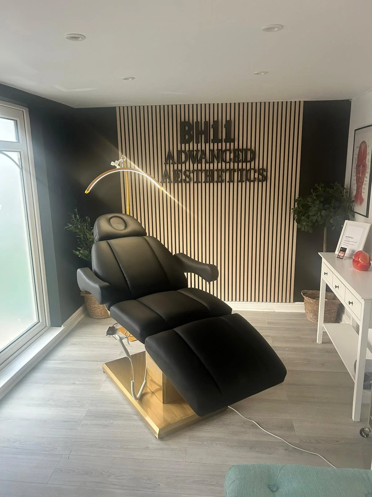 Interior of a skincare clinic or spa with a black treatment chair, a wooden base, and adjustable lamp, near a window with sheer curtains. The wall behind features a sign that reads 'BH11 Advanced Aesthetics' with decor including framed art and plants.