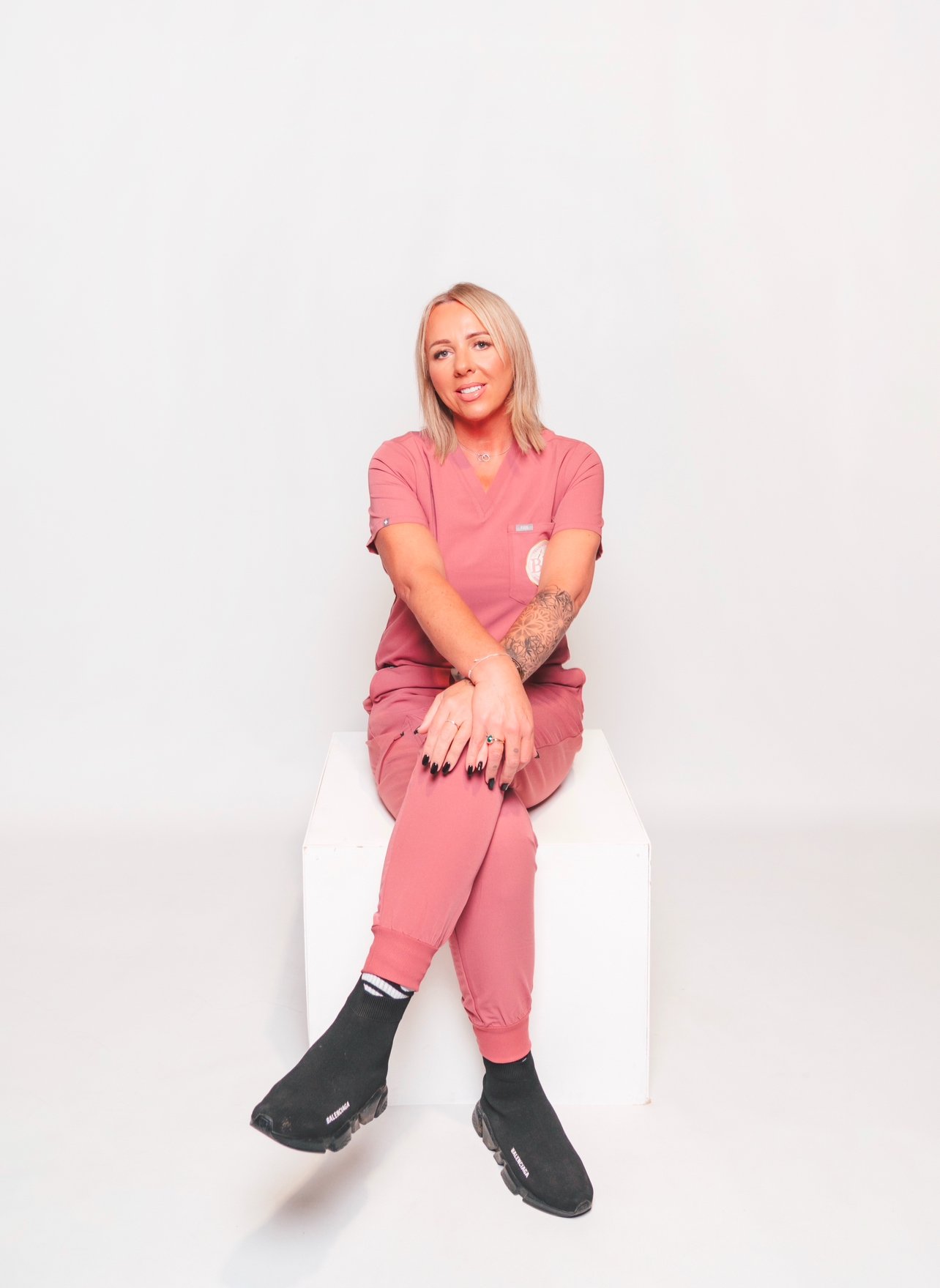 A woman in pink scrubs sitting on a white box against a white background, with tattoos on her arms and black slip-on shoes.