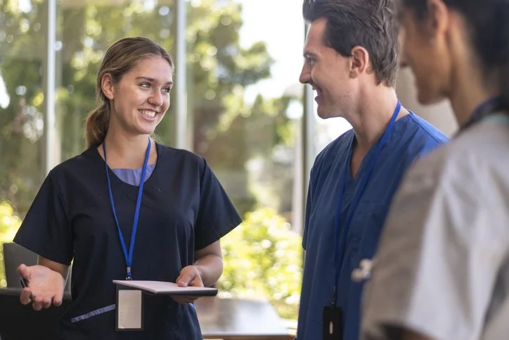 Three healthcare professionals having a conversation indoors, with sunlight and trees visible through the window behind them.