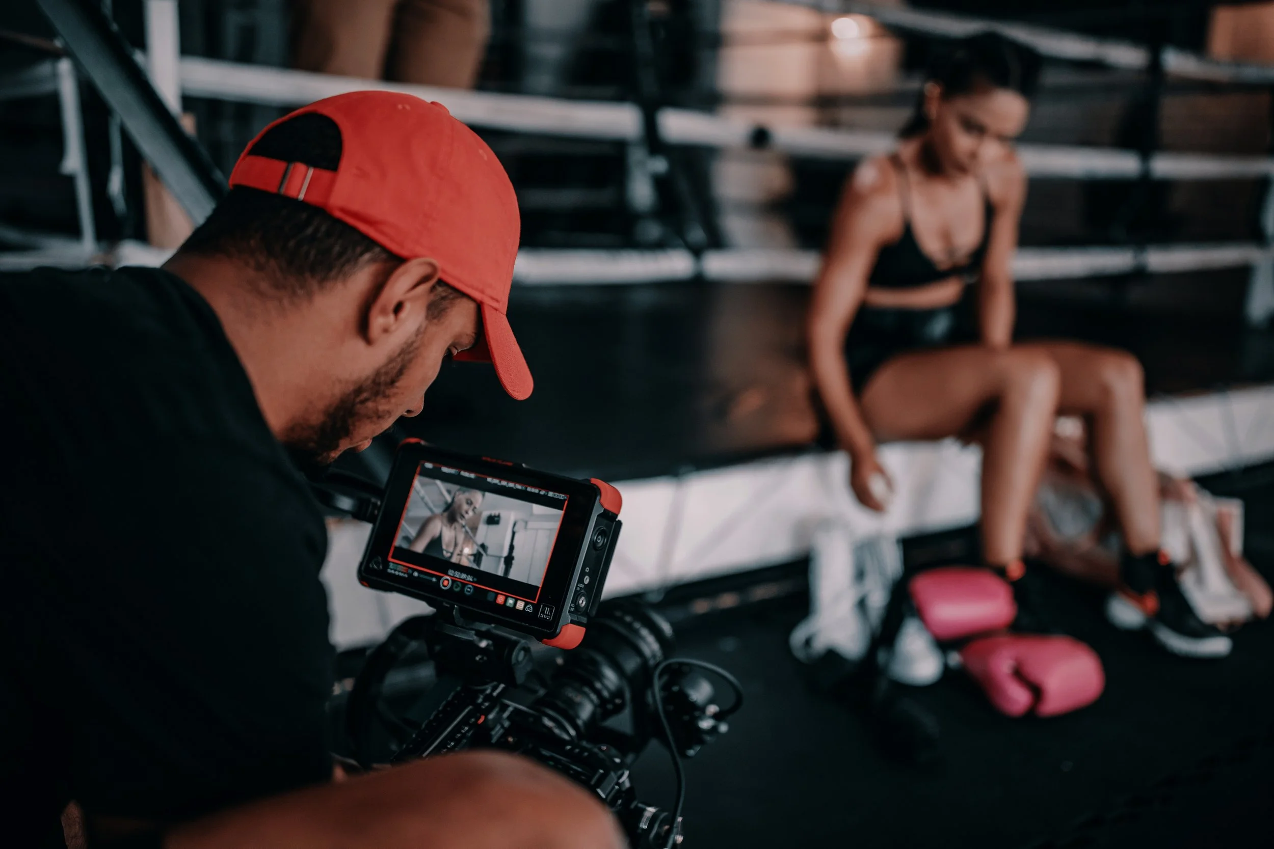 A man wearing a red cap operates a camera while filming a woman sitting on a boxing ring corner bench.